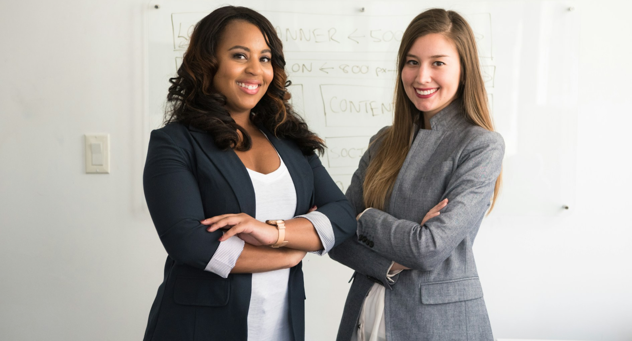 Two business women smiling with arms folded in front of a white board